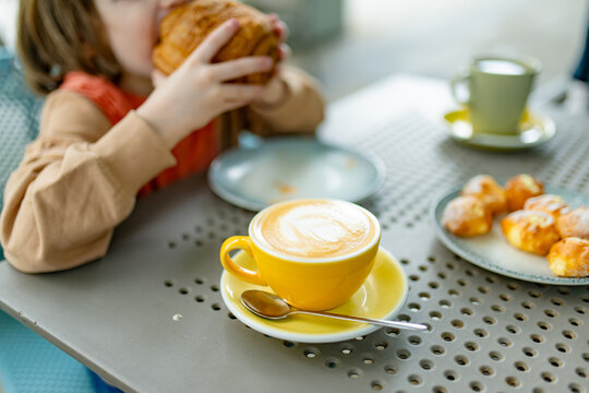 Young child eating a large pastry at a cafe while a parent enjoys a cup of coffee. Family outing illustrates a small moment of relaxation and self-care during everyday parenting.