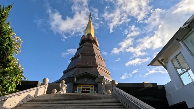 Wide shot of a large, dome-shaped Buddhist temple with a golden spire in northern Thailand, featuring many steps with railings leading up to it under a bright blue sky.