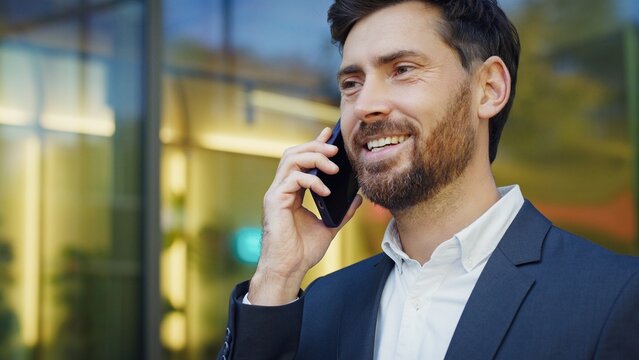 Caucasian businessman in formal suit talking on smartphone with cheerful expression. Describing good news from office to family near modern glass building. Positive attitude outdoors.