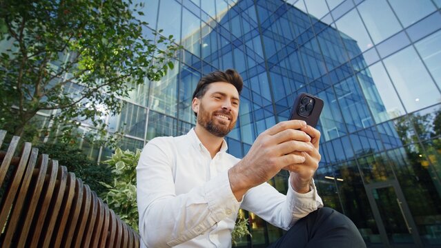 Smiling man in white shirt using smartphone while sitting on a park bench