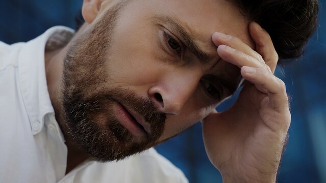Close up view of bearded Caucasian male touching forehead with hand showing stressed facial expression. Man in white shirt feeling worried and thinking deeply after unexpected bad news outdoors.
