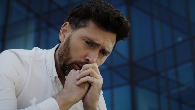 Upset Caucasian male in white shirt leaning forward clasping hands near face with worried expression. Thoughtful man sitting outside glass office building reflecting on difficult situation.