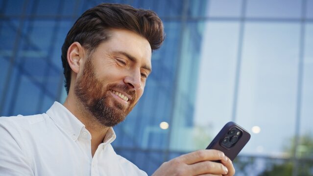 Side camera view of Caucasian male with beard wearing white shirt smiling while using smartphone. Man texting with someone on social media outside near glass modern business center.