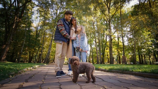 Happy Caucasian family standing together in green park smiling while spending weekend with brown fluffy dog. Parents and daughter enjoying relaxing outdoor walk under bright sunlight.