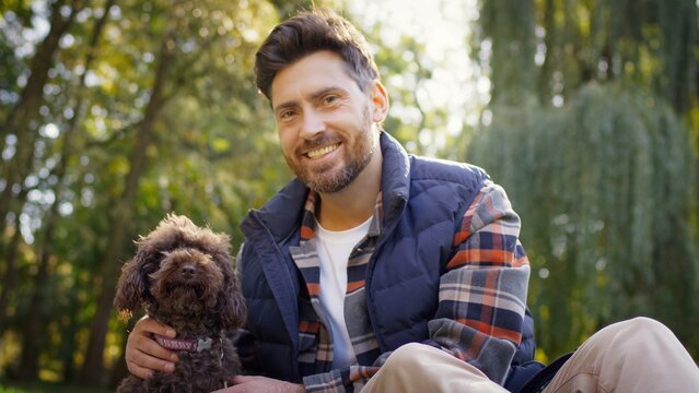 Smiling Caucasian man with beard sitting outdoors in park petting small brown dog. Handsome male in plaid shirt and vest enjoying relaxing sunny day. Sitting on ground together with pet.