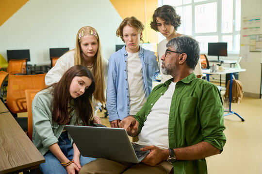 Indian middle aged male teacher explaining lesson on laptop to diverse group of teenagers in classroom. Image suitable for school marketing, digital learning, and modern education concepts