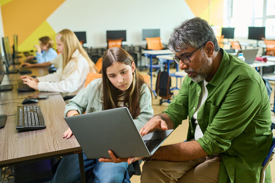Middle aged Indian male teacher helping Caucasian teenage girl with laptop in computer class. Image represents digital literacy and is ideal for school curriculum marketing