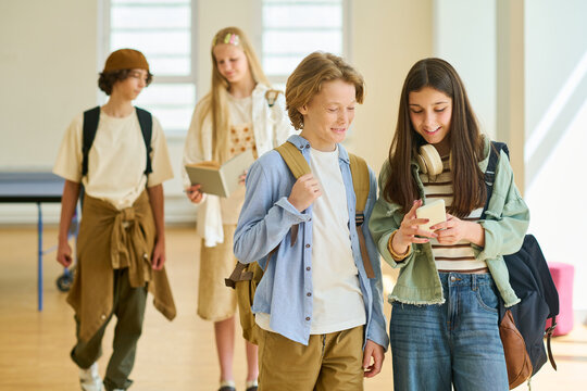 Caucasian teenagers walking in school hallway and looking at smartphone screen. Image represents digital lifestyle and is suitable for educational marketing campaigns