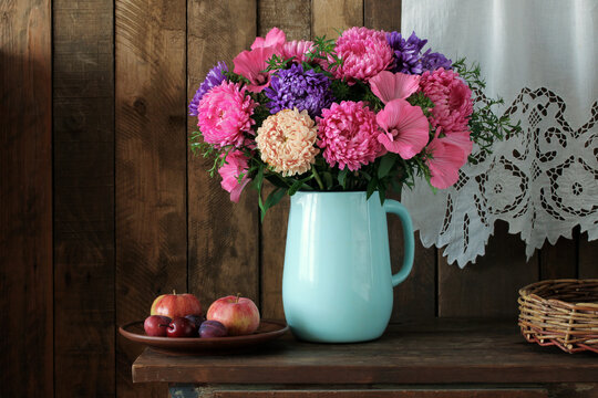 Garden flowers and fruits. A still life with asters and lavatera in an enamel jug, apples and plums on a plate.
