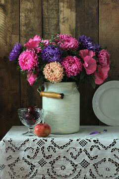 A rustic still life with asters and lavatera in an old can, apple and plums on a table with a lace tablecloth.