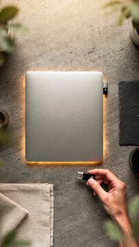 Flat lay top view of a minimalist modern workspace with a closed silver laptop backlit with warm light and a hand holding a USB flash drive
