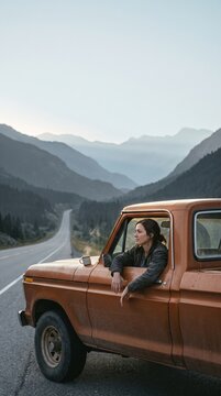 Young woman enjoying a morning coffee break in a vintage orange pickup truck during a scenic mountain road trip