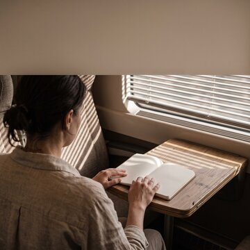 A woman reads a blank notebook while sitting at a wooden table next to a sunny train window during a peaceful trip
