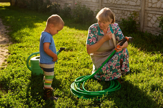 A child stands beside an adult in a garden. The adult is holding a hose and explaining something. Sunlight shines on the green grass. It is afternoon
