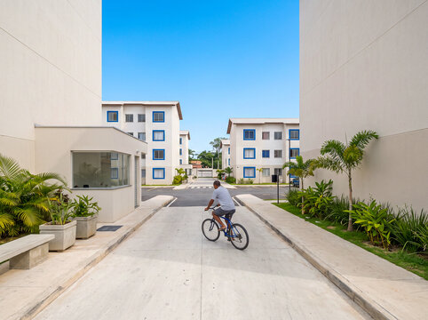 A man rides a bicycle through a modern gated residential community with white apartment buildings and tropical landscaping under a clear sky