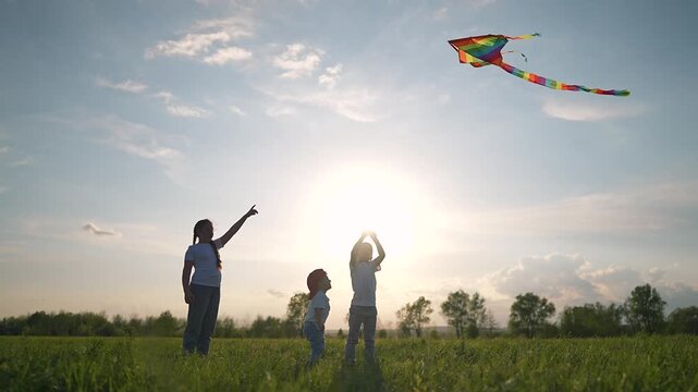 Group of children watch kite fly high. Boy holds string in field. Grass rustles under child feet. Sunset makes silhouette glow. Outdoors play with kite at sunset. Girl laughs near child in grass.