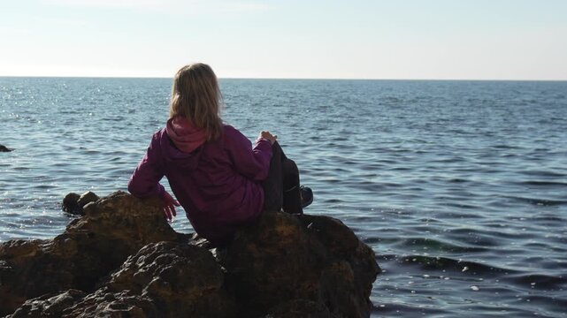 Woman, ocean, travel, sits on a sunny coastal rock, contemplating the vast blue seascape during a solo vacation trip