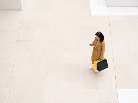 High Angle View of an Asian Woman in a Trench Coat Holding Shopping Bags and Using a Smartphone on Tiled Floor