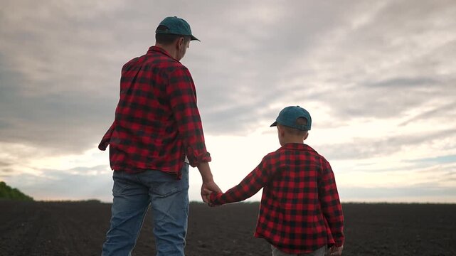 Boy father holding hands while walking through field. Outdoor view of nature surrounds them. Child walks calmly. Holding hand brings comfort. Field expands wide. Boy steps forward holding father hand.