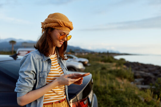 Cheerful woman outdoors, looking at her phone by a car, striped shirt, denim jacket, knit hat, golden hour glow, coastal landscape, relaxed lifestyle, natural colors