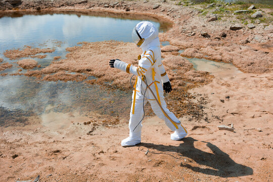 Woman astronaut in white spacesuit walking along the shore of an alien planet lake