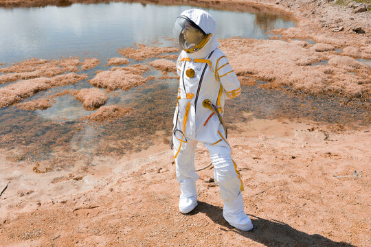 Astronaut standing by red lagoon on alien planet surface in white spacesuit, wide shot