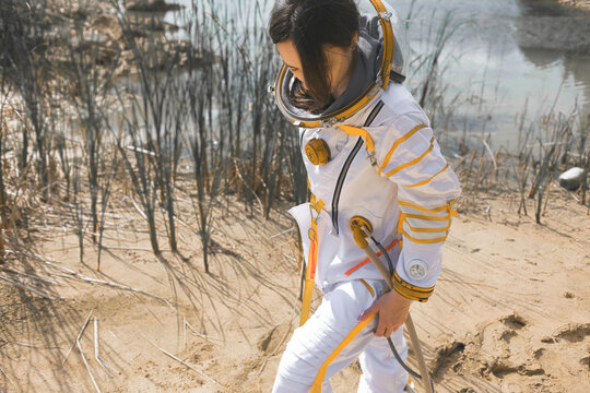 Woman in a space suit descending a sand slope toward water framed by dry reeds