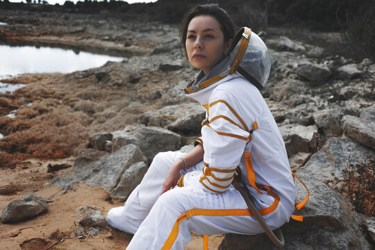 Serious woman astronaut looking at camera while sitting on a rock by an alien lake