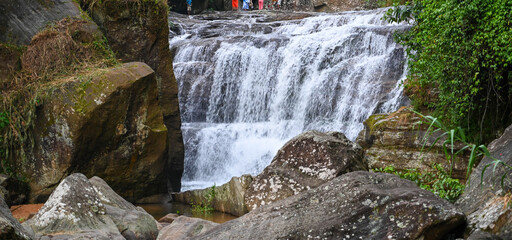 Panoramic view of a scenic waterfall cascading down rocky cliffs and large boulders surrounded by dense green forest foliage, ideal for natural landscape stock. © Fazlerabbi