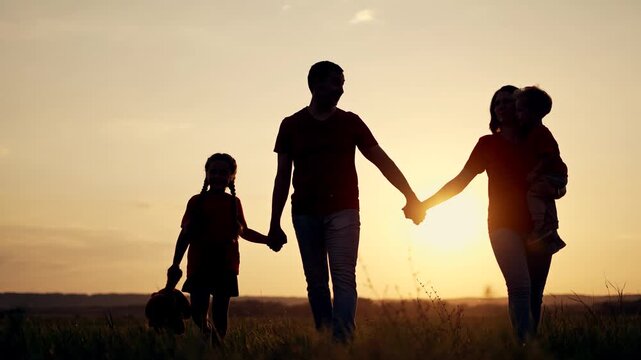 Happy family walks through field at sunset. Father and mother hold hands with children. Daughter carries teddy bear. Mother holds young son. Family silhouettes glow in sunset at golden hour.