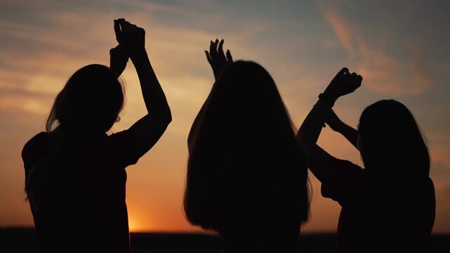 Three girls silhouettes dance with raised hands at sunset. Friends enjoy freedom outdoors. Women silhouettes celebrate together. Sunset sky glows orange. Happy friends dance in evening light.
