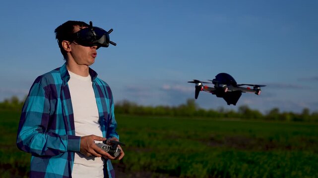 Man flying drone with FPV goggles outdoors. Pilot holds remote controller in green field. Drone hovers nearby during sunset. Man controls flying quadcopter. FPV pilot enjoys drone flight in nature.