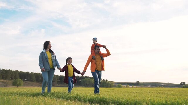 Happy family walking through green field at sunset. Parents and children hold hands outdoors. Father carries son on shoulders. Family walks together in countryside. Happy kids enjoy nature walk.