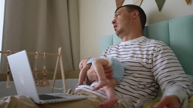 Young, tired father trying to multitask, working on a laptop while holding his newborn baby. The freelancer dad is exhausted, yawning and falling asleep from lack of rest in the bedroom