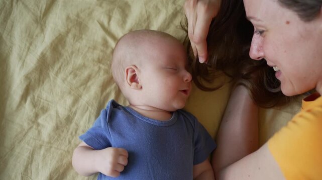 Young mother affectionately plays with her infant son, tickling his cheek with her hair as they lie face to face on a bed. The newborn boy reacts with contentment to his mom's touch