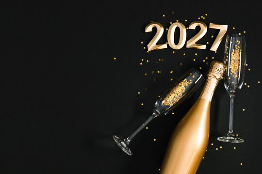 Happy New Year 2027 Elegant Background. Gold Champagne Bottle, Flutes and Star Confetti on Black. Festive Holiday Flat Lay Layout with Left Side Copy Space for Greeting Card or Banner Design