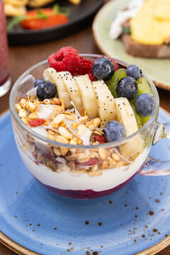 layered yogurt bowl served in clear cup with bananas, kiwis, raspberries, blueberries, chia seeds, and granola on a blue ceramic plate