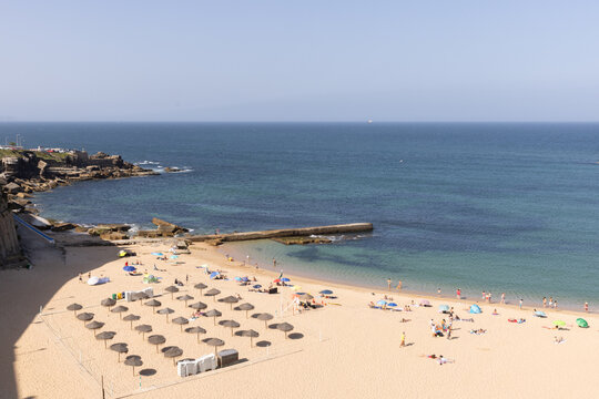 afternoon sunny blue sky wide shot of Ericeira coast in Portugal with cliffs, ocean, and colorful beach goers and umbrellas