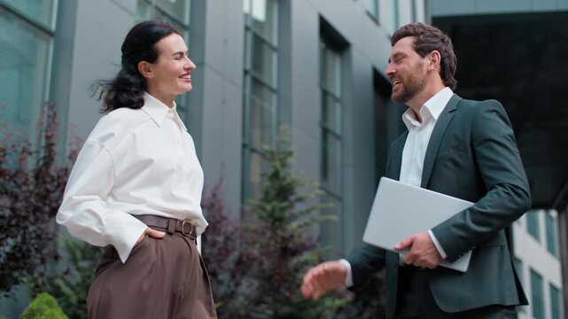 Two Caucasian mature business colleagues giving high five talking in city outside office. Confident businessman with businesswoman talk outdoors man with woman discussing team successful working deal