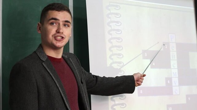 A young male teacher standing by a chalkboard and projection screen, pointing with a pointer at a crossword puzzle during a lesson. a smart educator mapping out information in a classroom