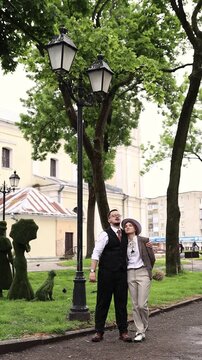 An eccentric couple standing on a park path near a vintage lamppost, with the groom smoking a cigar. Groom smoking a cigar, unique vintage bride fashion, nonconformist wedding concept, and elegance