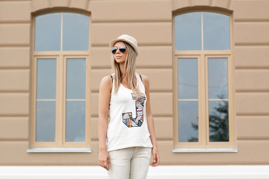 Stylish blonde woman in aviator sunglasses, fedora hat, and graphic tank top standing outdoors near a modern building facade. Trendy urban street fashion look for summer vacation and lifestyle.