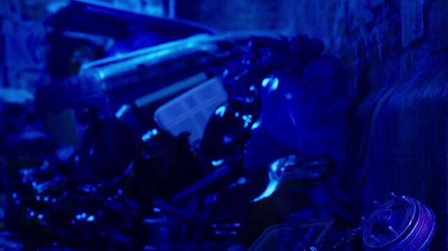 A collection of broken objects is stacked in a narrow alley. The scene is lit with blue light, creating shadows and highlighting the shapes of the discarded materials.