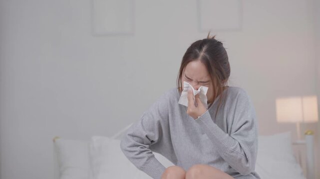 	
Cold, sickness and flu symptoms concept. Young Asian woman sneezes into a tissue while sitting on a bed at home, feeling unwell. Cold, sneezing, sickness, flu, healthcare