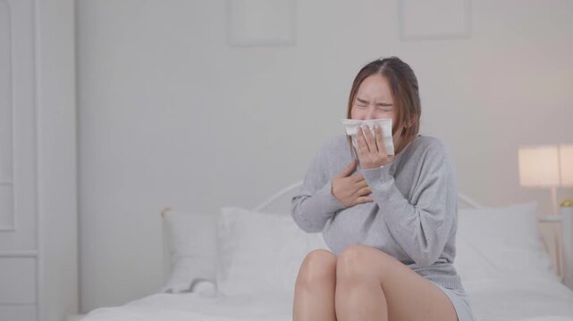 	
Cold, sickness and flu symptoms concept. Young Asian woman sneezes into a tissue while sitting on a bed at home, feeling unwell. Cold, sneezing, sickness, flu, healthcare