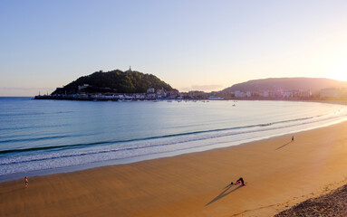 Fototapeta premium Couple walking at sunrise in La Concha Bay, San Sebastian Donostia, Basque Country, Spain. Scenic view of Urgull mountain and city skyline.