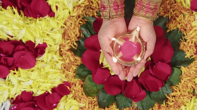 Overhead shot of Indian woman hands holding a lit diya oil lamp over a colorful flower rangoli pattern during Diwali festival celebration.