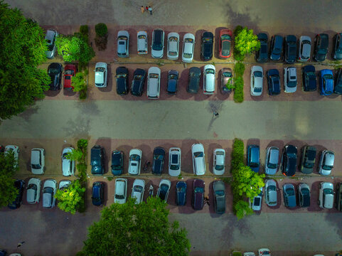 Aerial view of a busy parking lot filled with various cars parked in organized rows, surrounded by green trees and paved walkways in Gurgaon, Haryana, India.