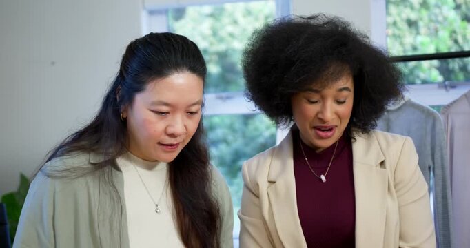 Diverse female team seeing swatch leaning forward pointing at tablet reviewing outfits in studio