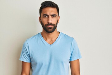 Obraz premium Young man with beard expresses doubt and distrust. His raised eyebrow and serious look show uncertainty. He stands indoors against a light gray background.